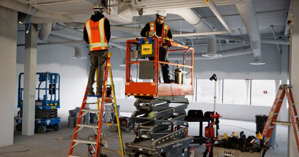two construction workers fixing HVAC on jobsite--one on a ladder, one on a lift.